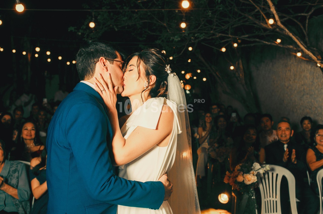 Un couple s'embrasse lors d'une cérémonie de mariage en extérieur, entouré d'invités et de guirlandes lumineuses.