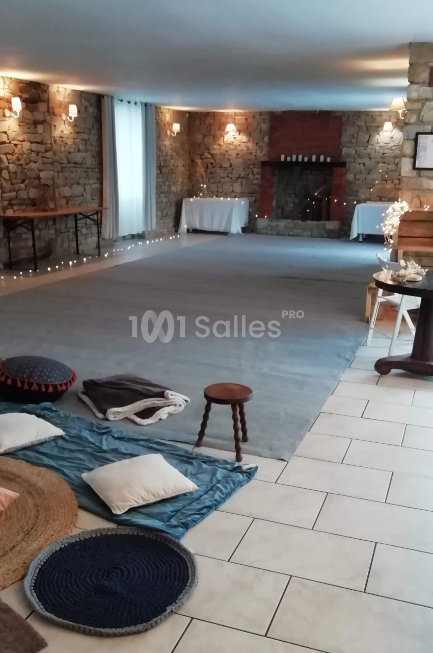 Salle lumineuse avec murs en pierre, tapis, coussins au sol, tables en fond et cheminée décorative.