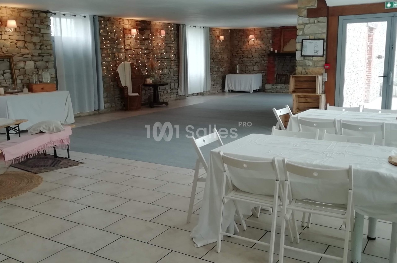 Salle lumineuse avec murs en pierre, tables blanches, chaises pliantes et espace dégagé pour des activités ou événements.