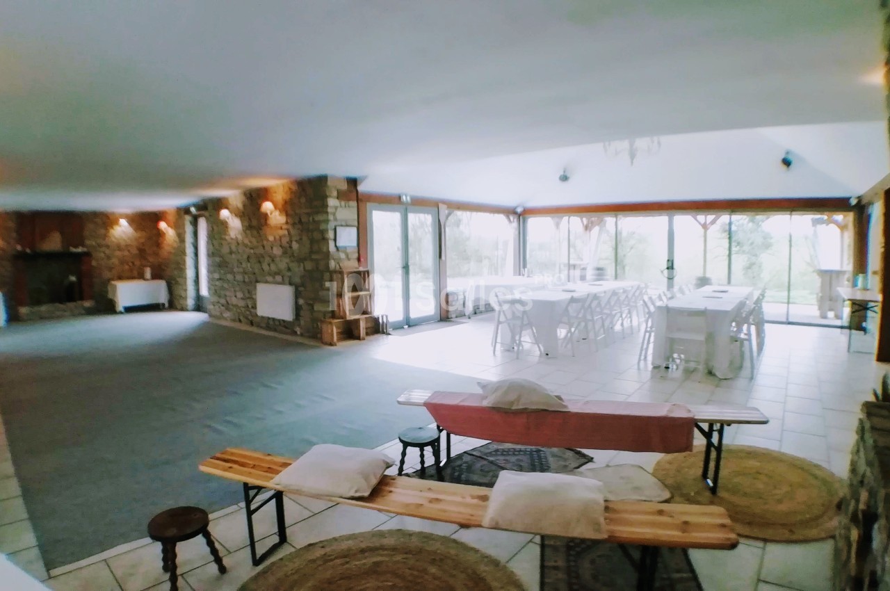 Salle spacieuse avec tables dressées, grandes baies vitrées, murs en pierre et espace aménagé avec bancs et coussins.