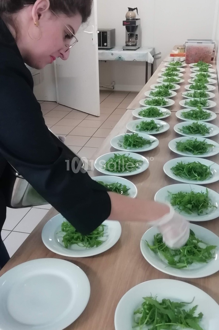 Une personne dispose des assiettes garnies de salade verte sur une longue table dans une cuisine.