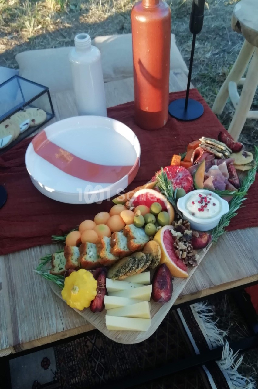 Plateau de charcuterie et fruits avec fromages, dips et garnitures sur une table en bois, décorée pour un repas en extérieur.