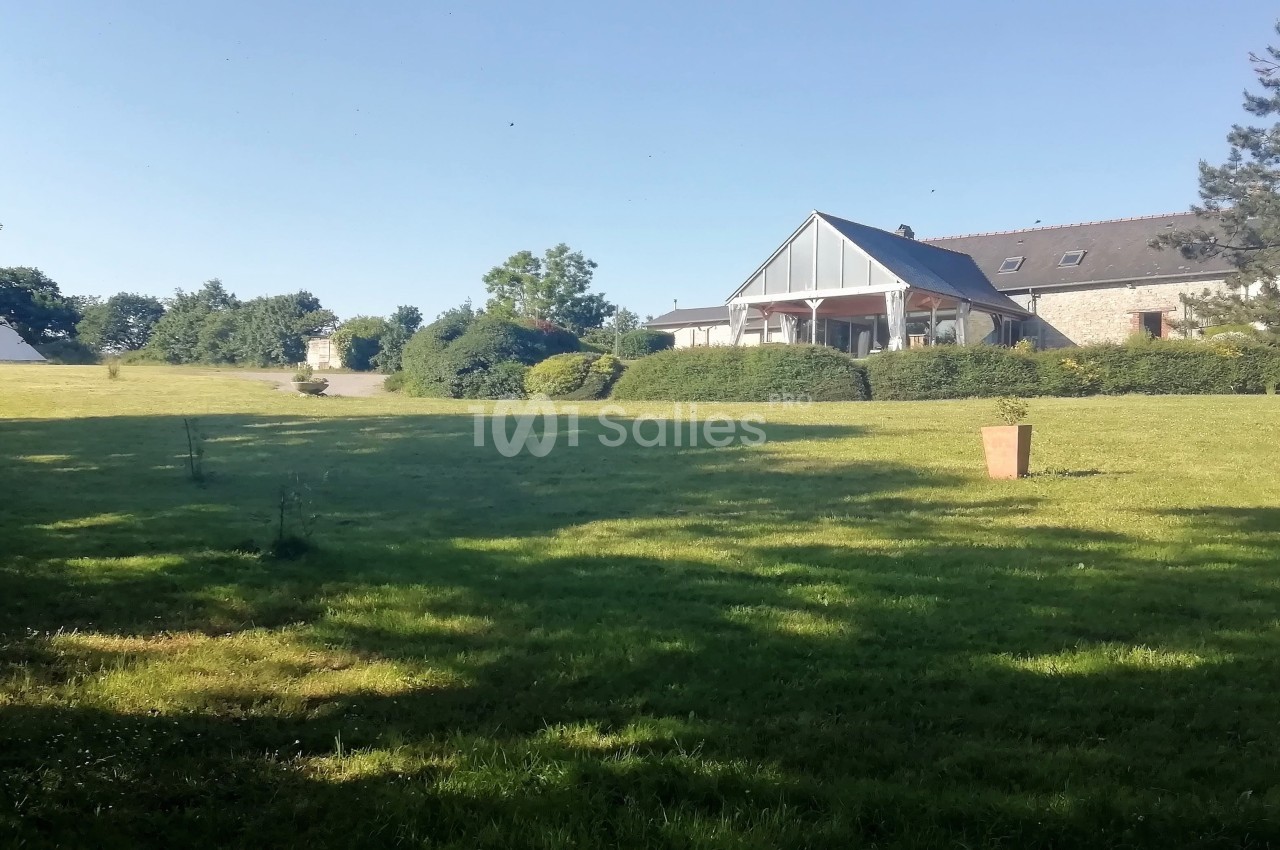 Grande pelouse verdoyante avec une maison en pierre et un toit en ardoise entourée de haies et d'arbres.