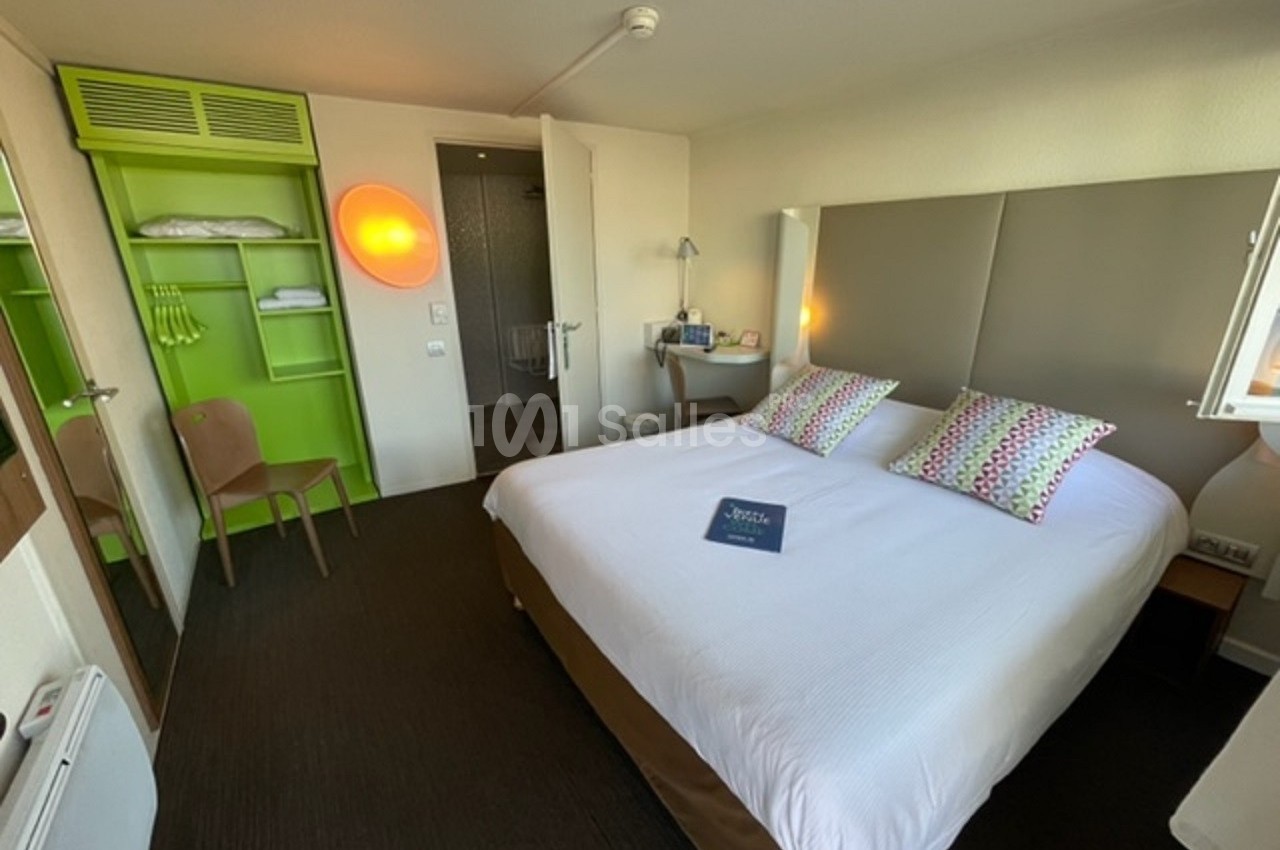 Chambre d'hôtel avec lit double, tête de lit rembourrée, étagères vertes ouvertes et salle de bain visible en arrière-plan.