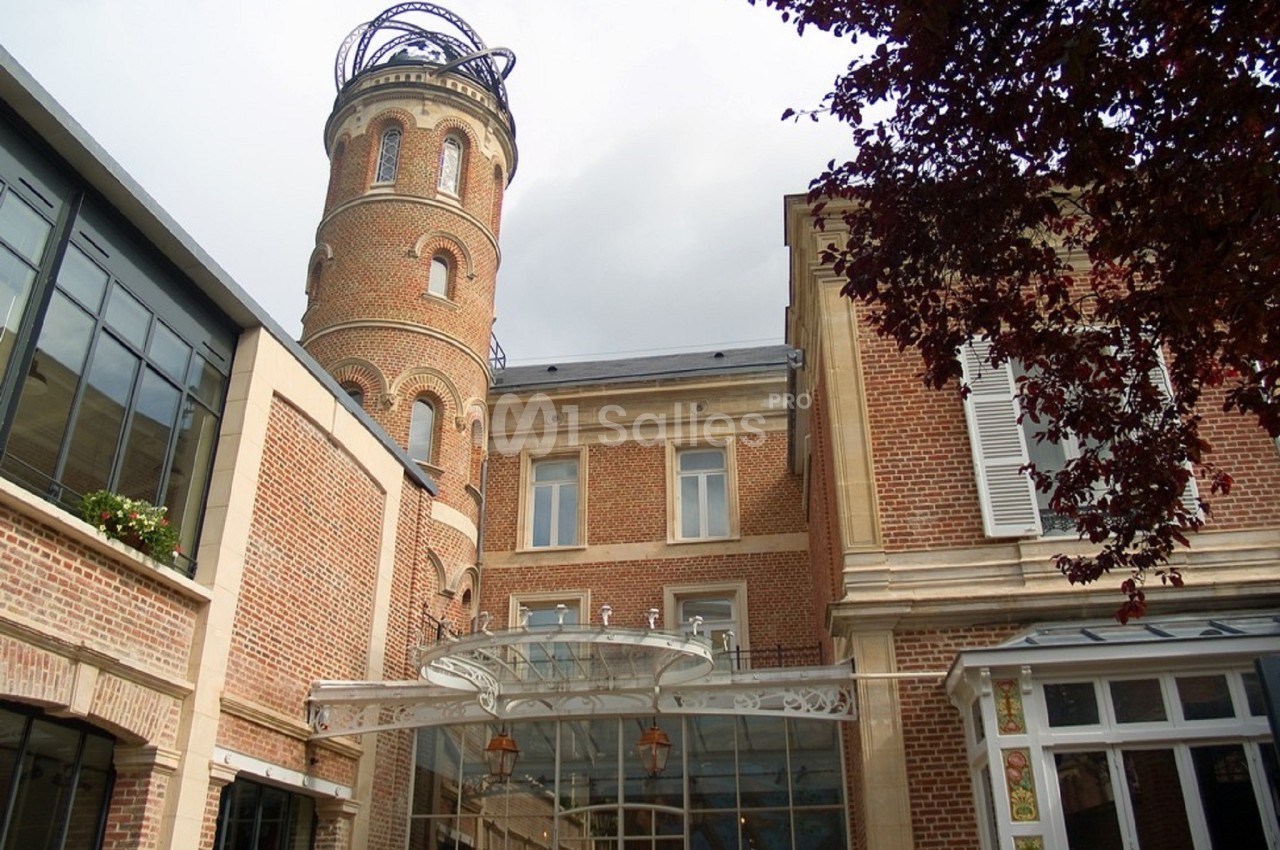 Bâtiment en briques rouges avec une tour cylindrique et une verrière, entouré d'arbres et d'éléments modernes.