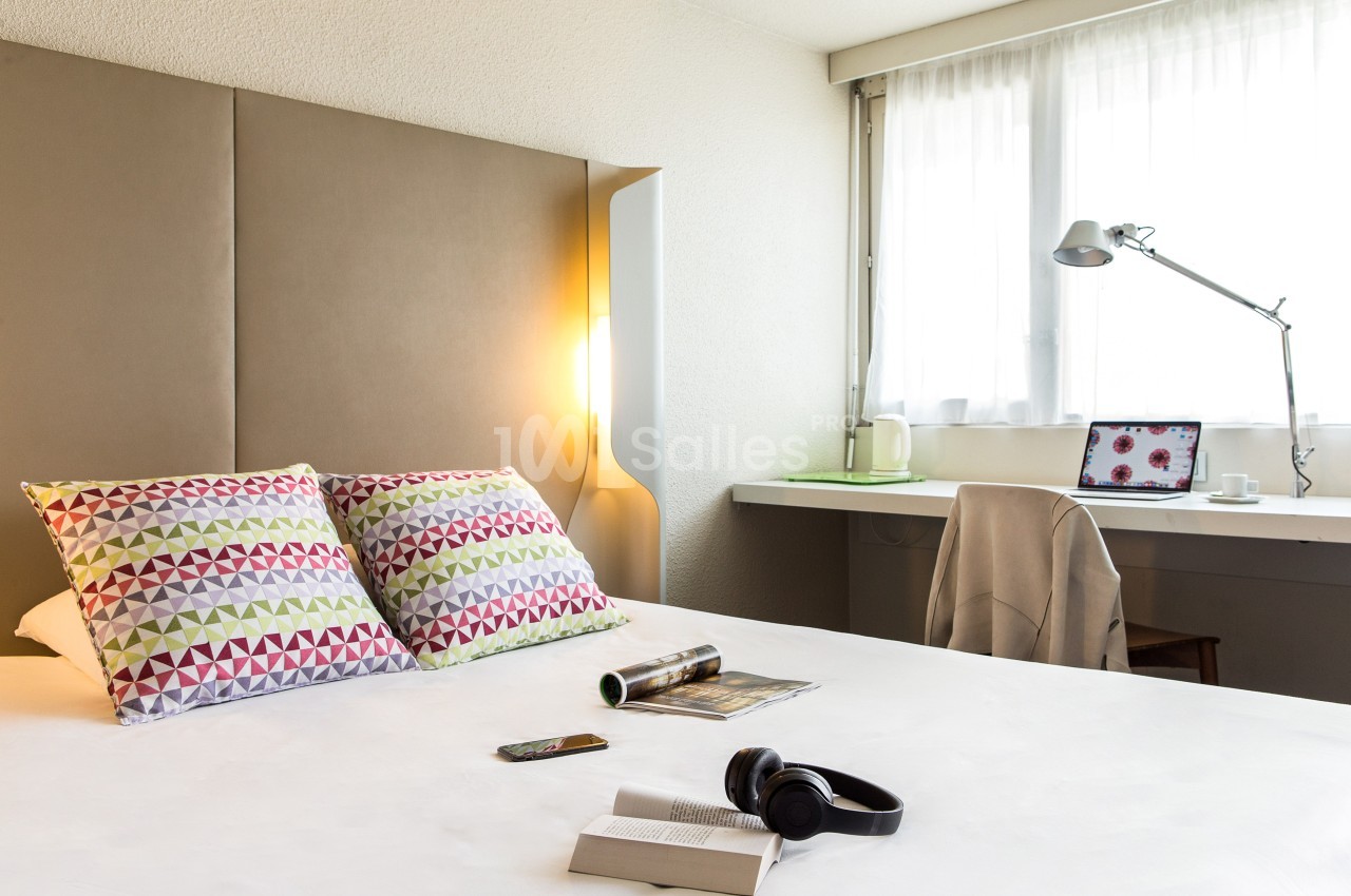 Chambre lumineuse avec lit, coussins colorés, bureau équipé d'une lampe, ordinateur portable et accessoires.