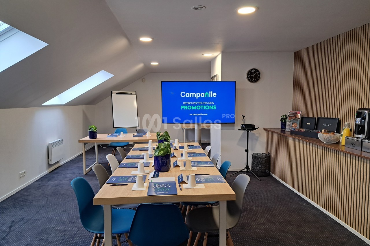 Salle de réunion lumineuse avec table rectangulaire, chaises colorées, écran, paperboard et coin café en arrière-plan.