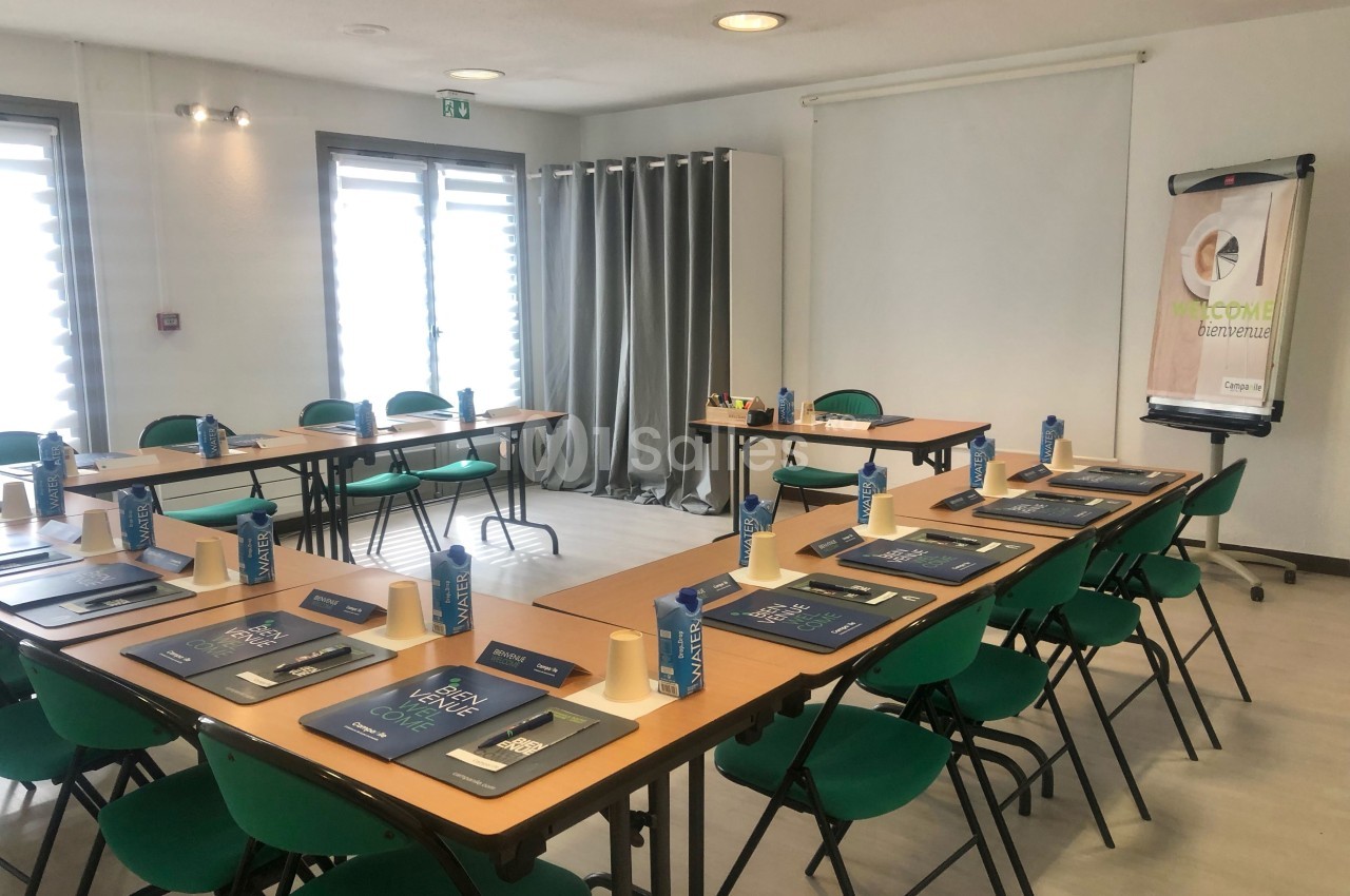Salle de réunion lumineuse avec tables disposées en U, chaises vertes et matériel de conférence installé.