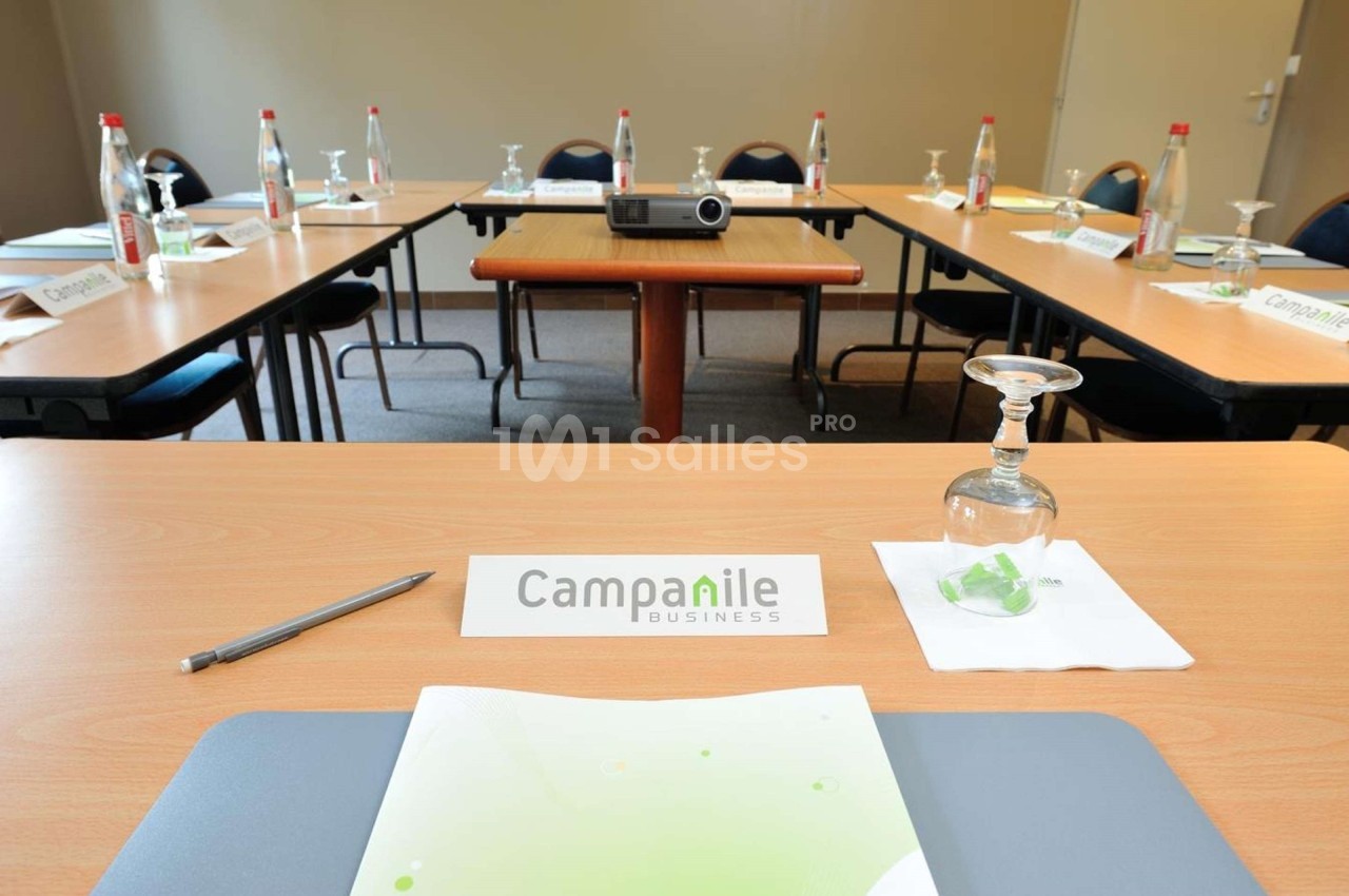 Salle de réunion avec tables disposées en U, équipée de bouteilles d'eau, verres, bloc-notes et un projecteur.