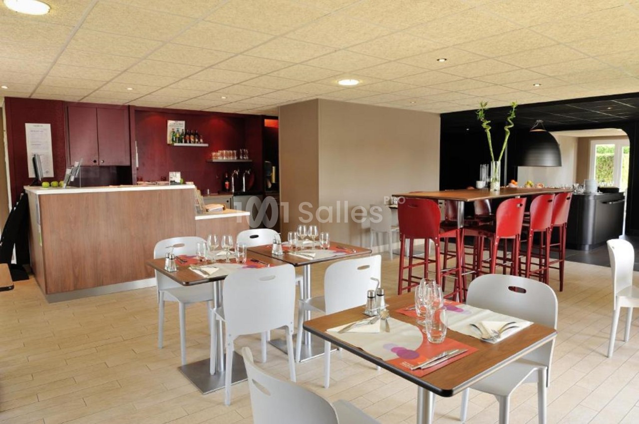Salle de restaurant lumineuse avec tables dressées, chaises blanches et rouges, et un comptoir en bois au fond.