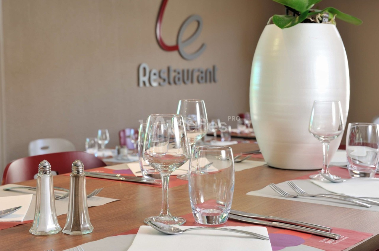 Table dressée avec verres, couverts et serviettes, devant un mur beige avec un logo et une plante en pot blanc.