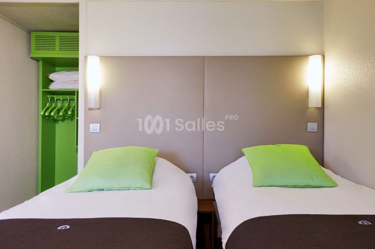 Deux lits simples avec tête de lit beige, éclairage mural et armoire ouverte avec cintres verts.
