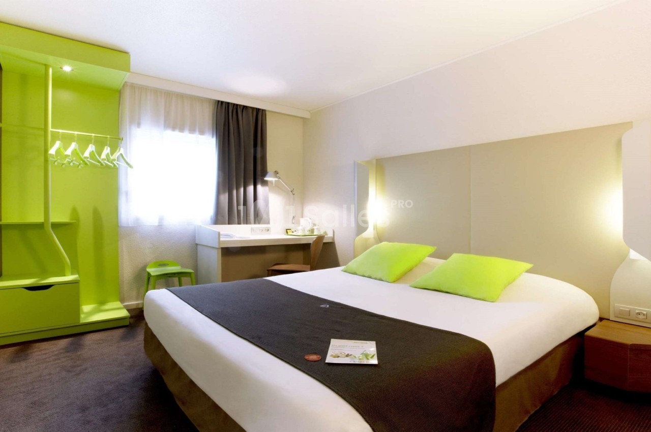 Chambre d'hôtel lumineuse avec lit double, bureau, armoire verte ouverte et fenêtre avec rideaux.
