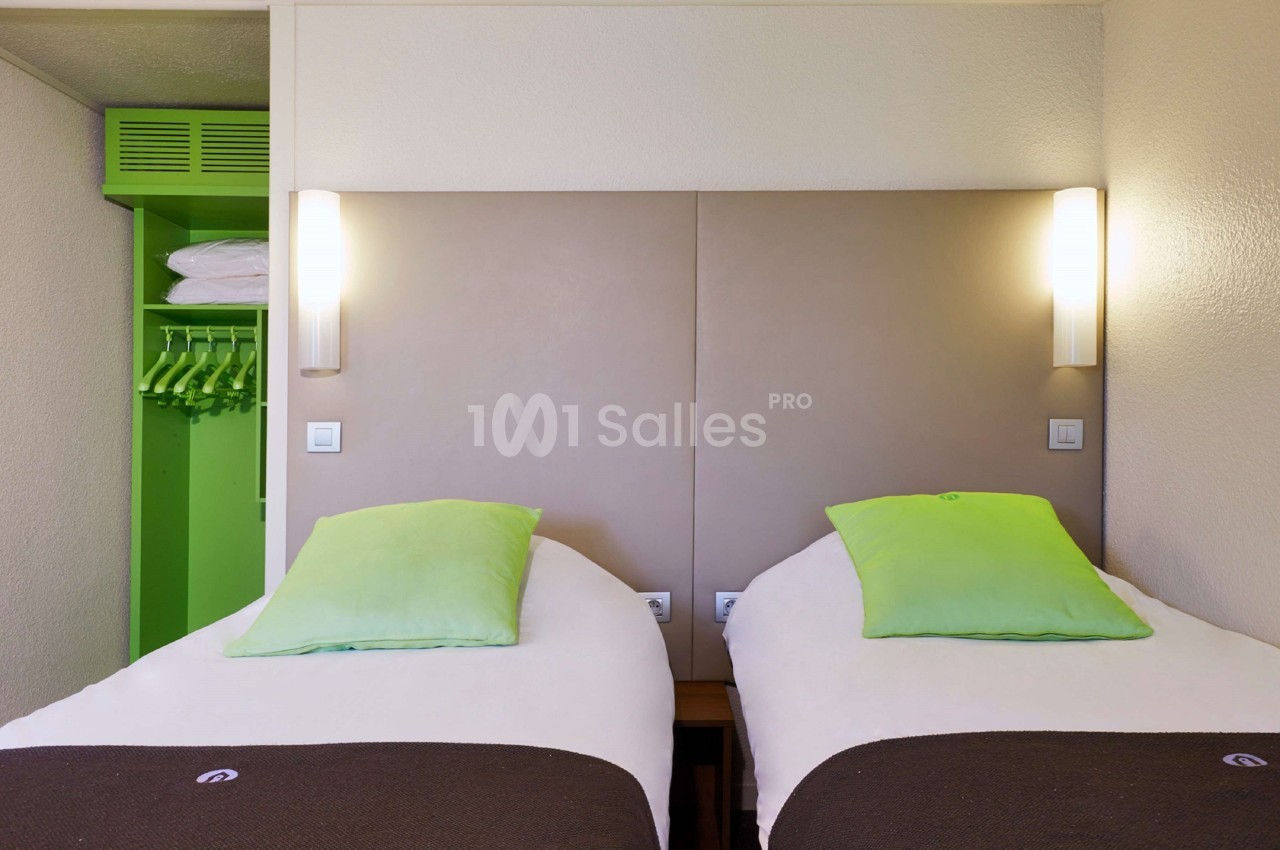 Deux lits simples avec têtes de lit beige, coussins verts, éclairage mural et armoire ouverte avec cintres.