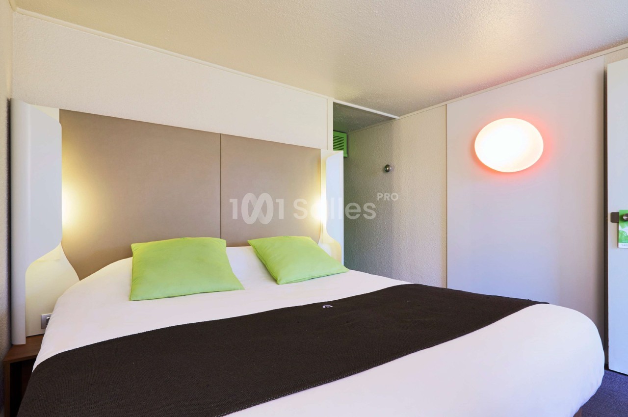 Lit double avec tête de lit beige, deux coussins verts, couverture sombre et éclairage mural dans une chambre lumineuse.