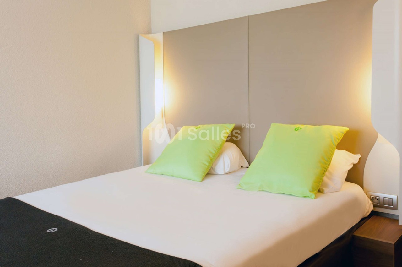 Lit double avec tête de lit rembourrée, deux coussins verts et éclairage mural intégré dans une chambre lumineuse.