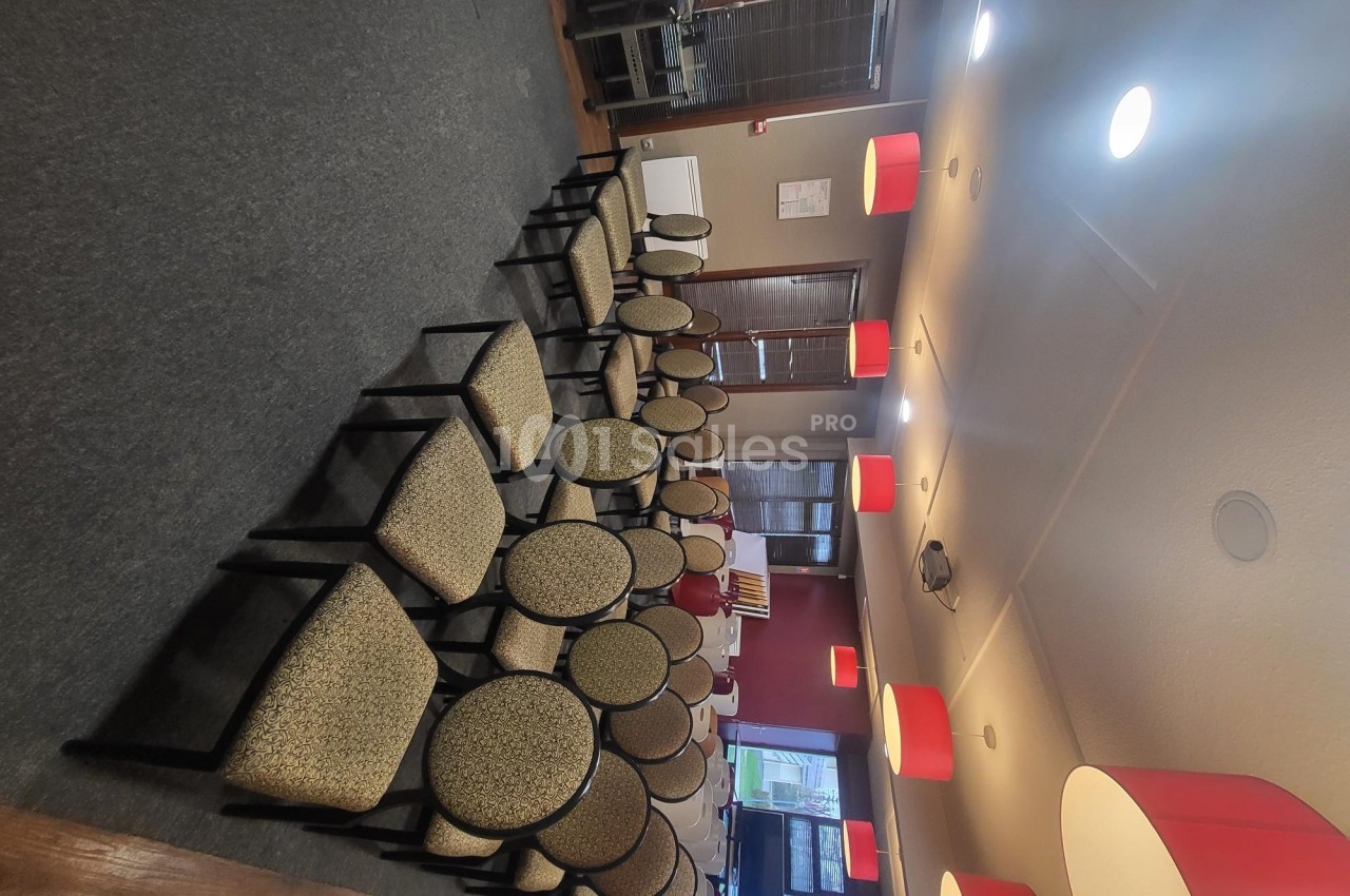 Salle de réunion avec des rangées de chaises rembourrées alignées, éclairée par des suspensions rouges.