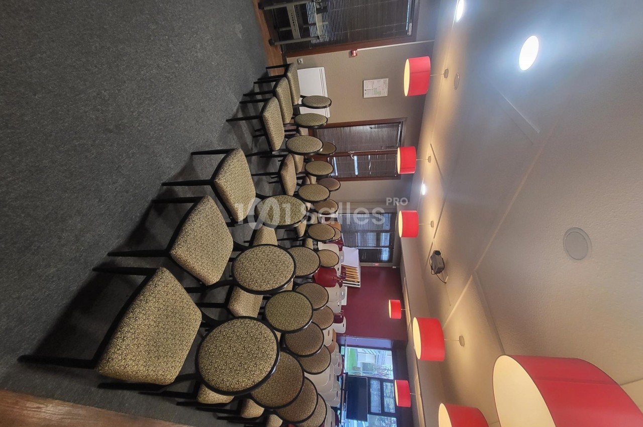 Rangées de chaises alignées dans une salle éclairée par des suspensions rouges, avec des fenêtres en arrière-plan.
