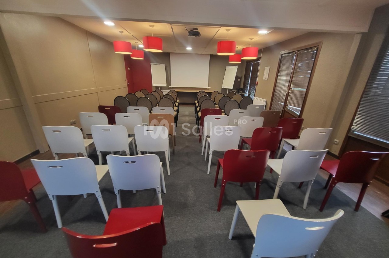Salle de conférence avec des rangées de chaises blanches et rouges face à un écran de projection et deux tableaux blancs.
