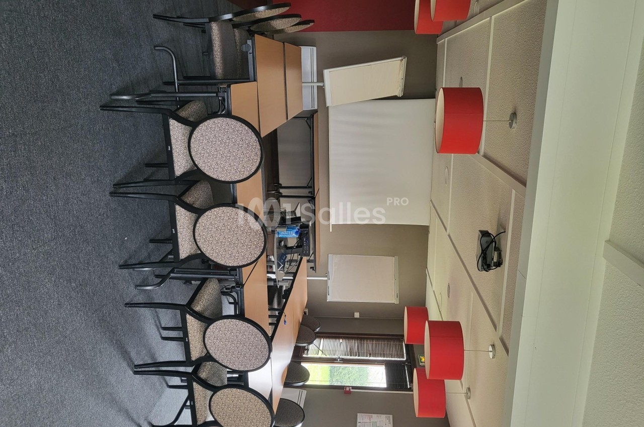 Salle de réunion avec chaises empilées, tables disposées en U, écran de projection et luminaires rouges.