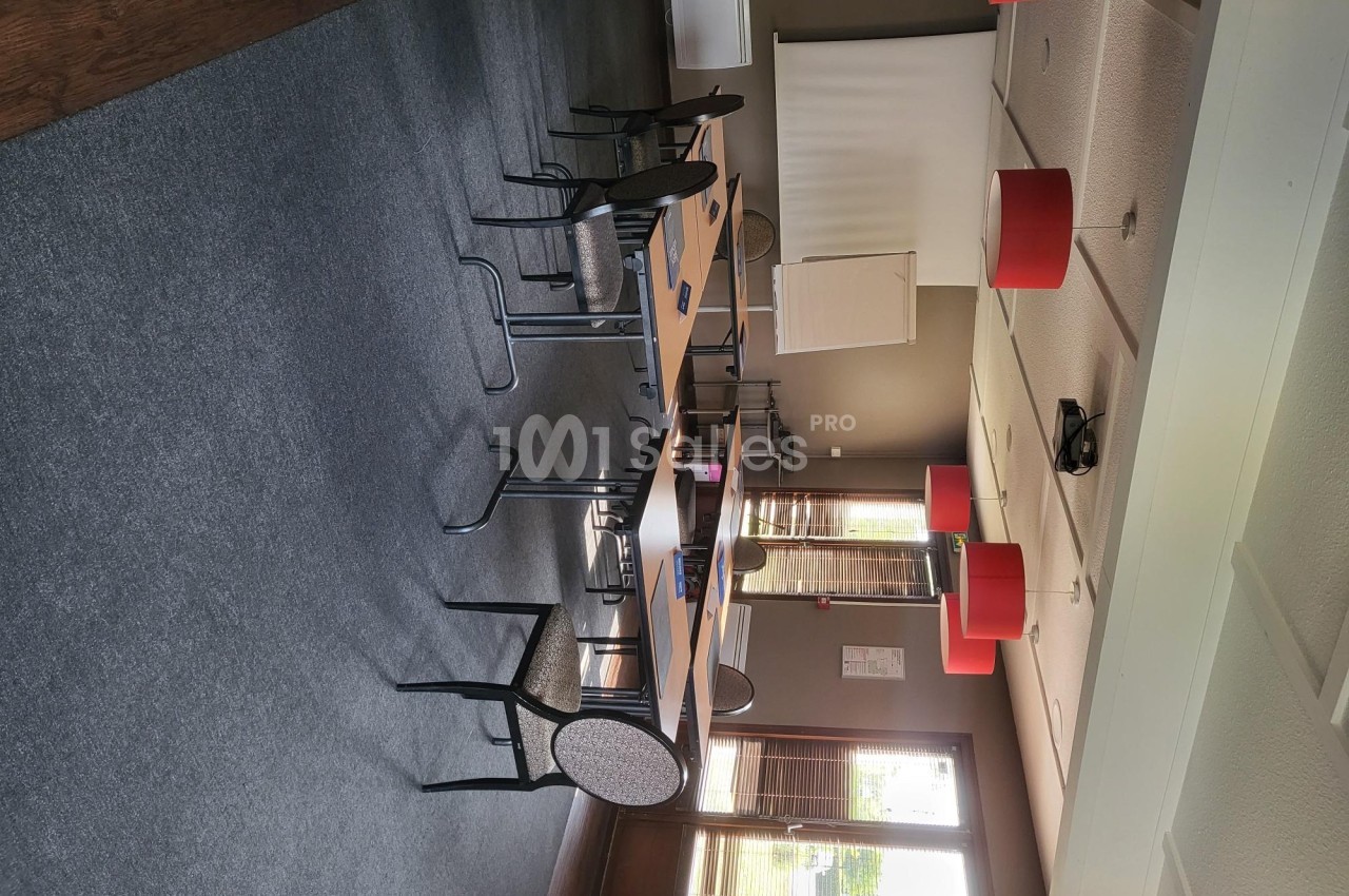 Salle de réunion lumineuse avec tables en bois, chaises, écran de projection et suspensions rouges au plafond.