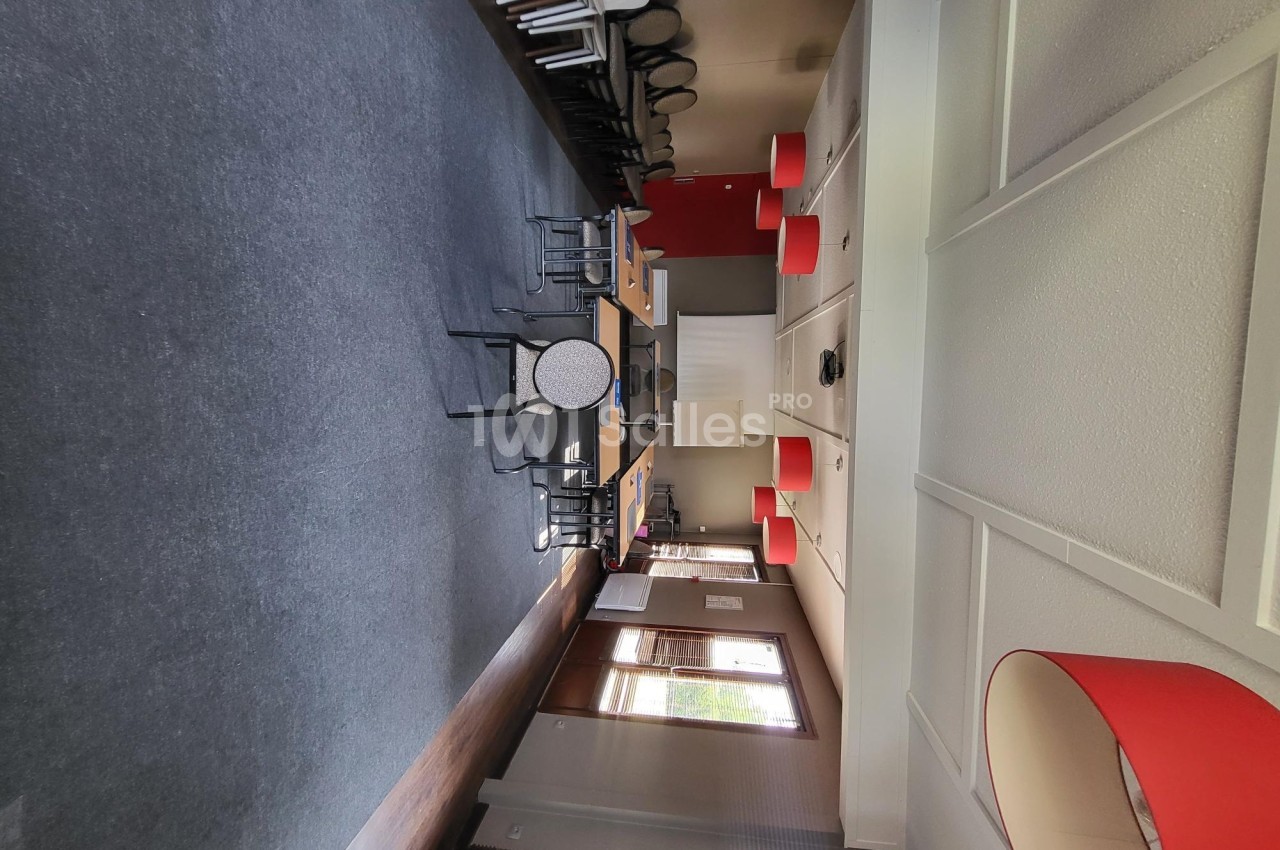 Salle de réunion lumineuse avec tables, chaises, murs clairs et lampes rouges suspendues.