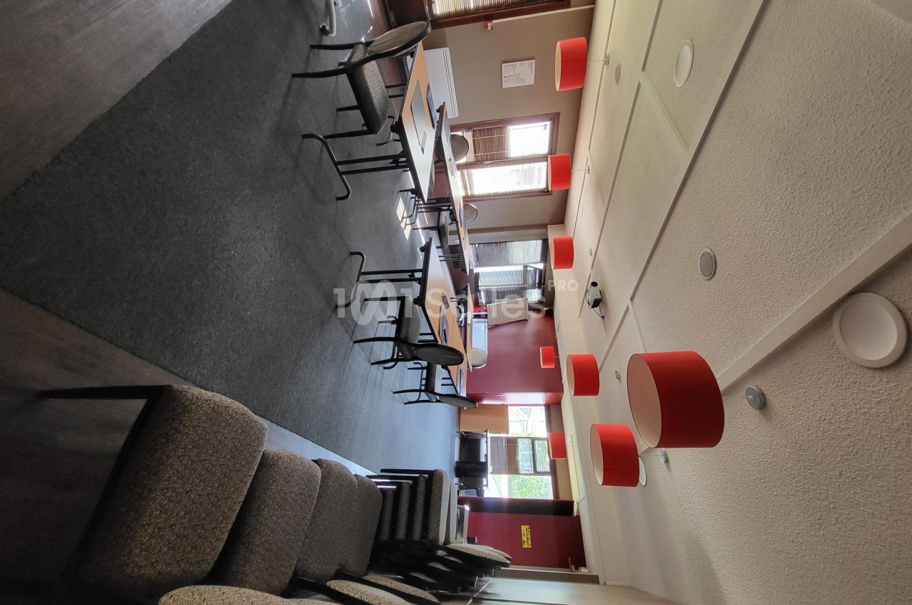 Salle de réunion avec chaises empilées, tables disposées en rangées et luminaires rouges suspendus.