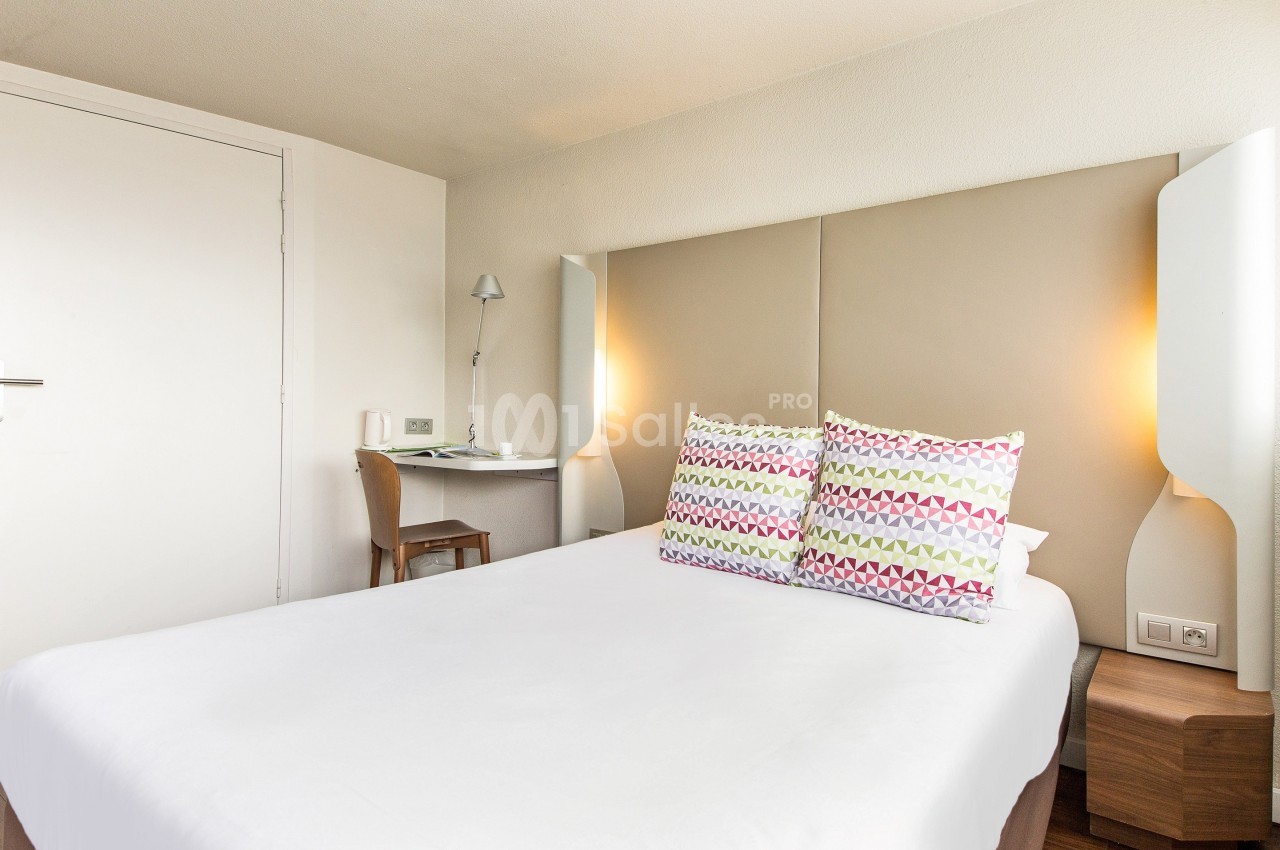 Chambre lumineuse avec un lit double, deux oreillers colorés, un bureau en bois et une lampe murale.