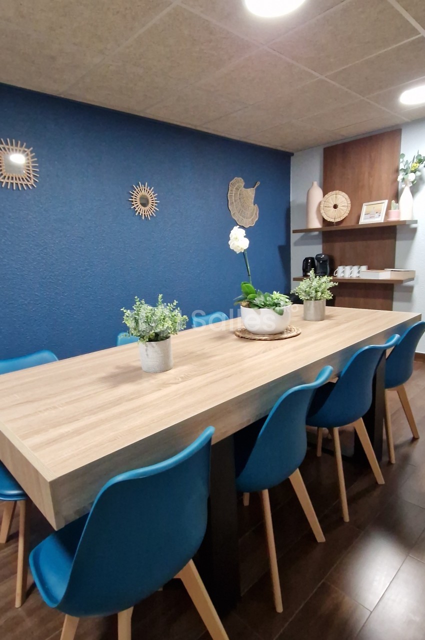 Salle à manger avec une grande table en bois, chaises bleues, mur bleu décoré et étagères en bois dans le fond.