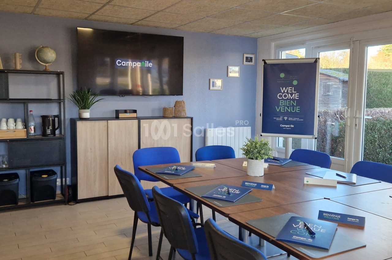 Salle de réunion lumineuse avec table, chaises bleues, écran mural, étagère et matériel de présentation.