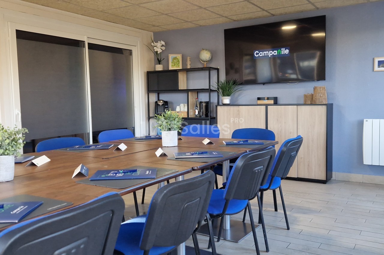 Salle de réunion lumineuse avec table en bois, chaises bleues, écran mural et affichage d'accueil.