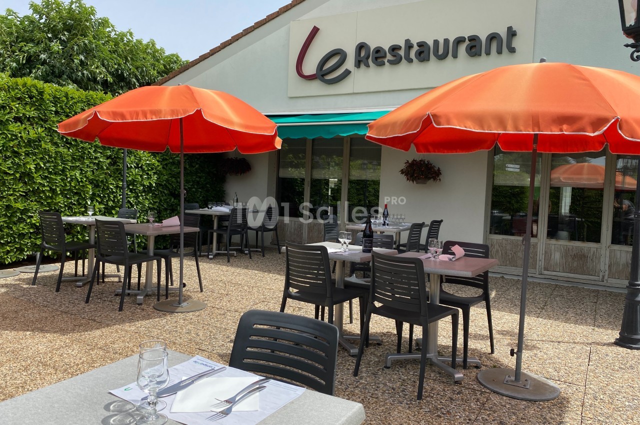 Terrasse d'un restaurant avec tables dressées, chaises noires et parasols rouges, devant une façade blanche.