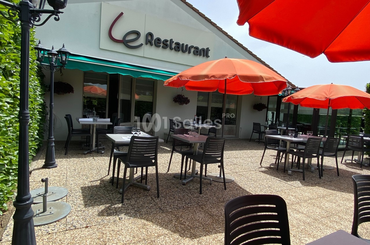 Terrasse d'un restaurant avec tables et chaises sous des parasols rouges, devant un bâtiment blanc.