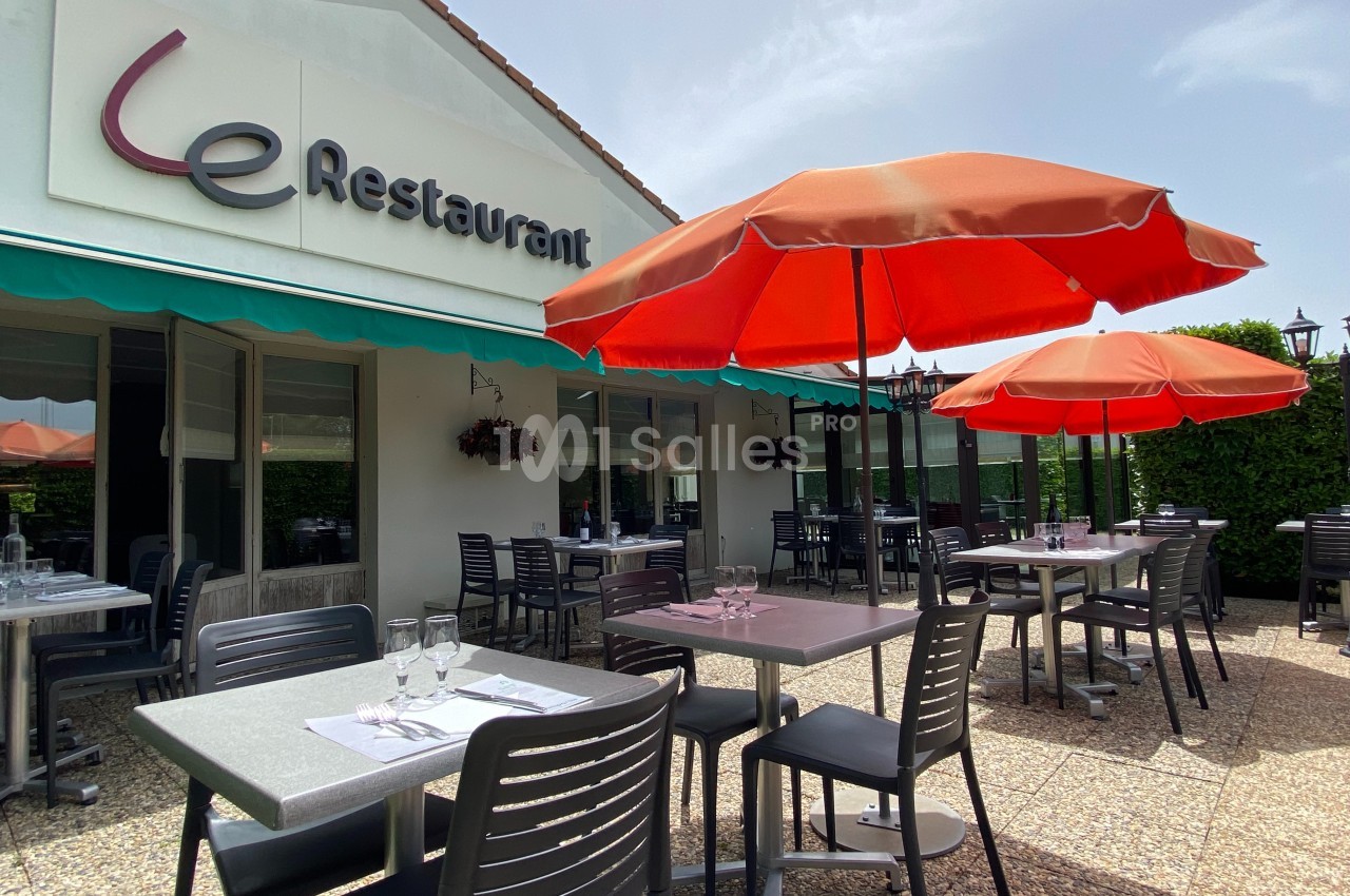 Terrasse d'un restaurant avec tables dressées, chaises noires et parasols rouges, devant une façade blanche.