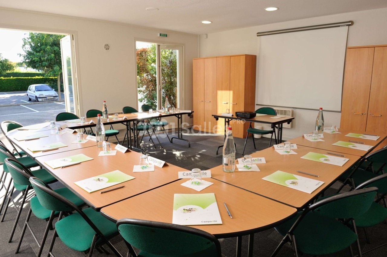 Salle de réunion lumineuse avec tables disposées en U, chaises vertes, documents, bouteilles d'eau et vidéoprojecteur.