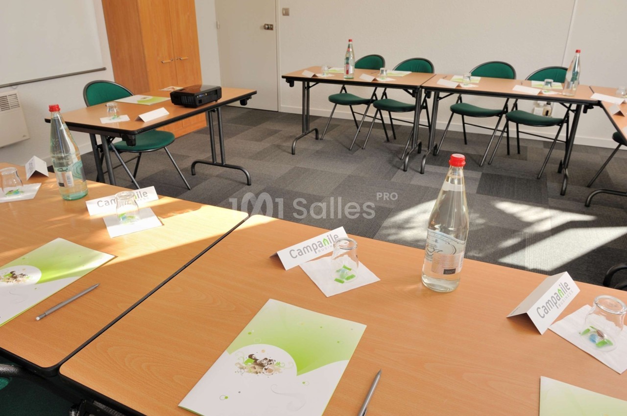 Salle de réunion lumineuse avec tables en U, chaises vertes, documents, bouteilles d'eau et matériel de présentation.