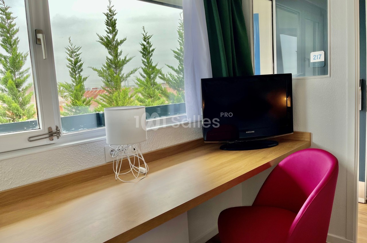 Bureau en bois clair avec lampe blanche, chaise rose, télévision et vue sur des arbres à travers une fenêtre.