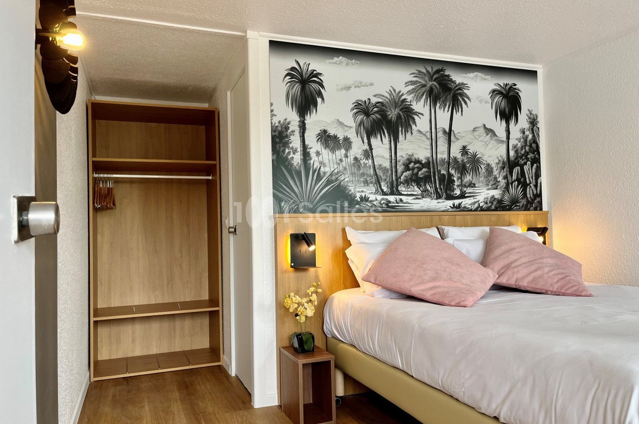 Chambre lumineuse avec lit double, tête de lit décorée d'une fresque tropicale, armoire ouverte et sol en bois.