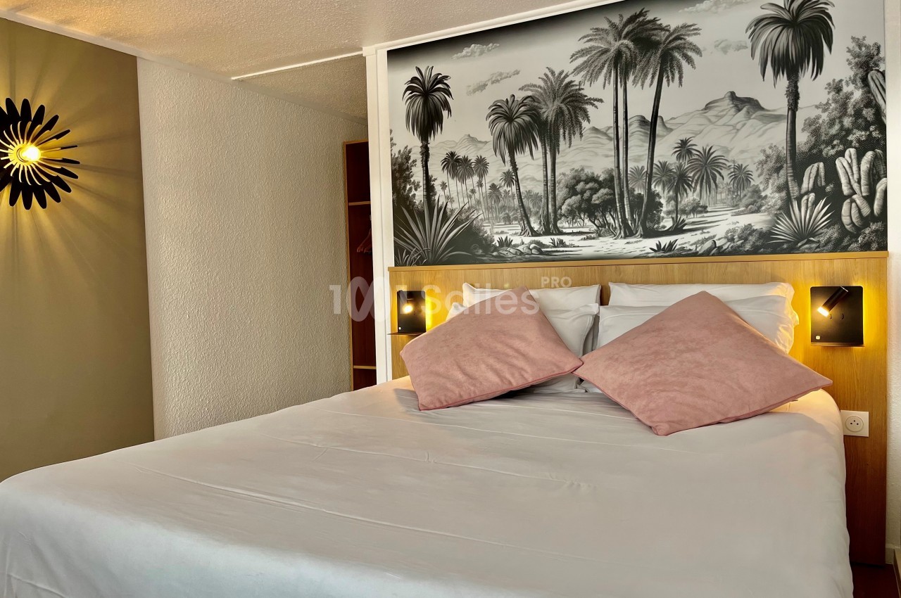 Lit double avec coussins roses, éclairé par des appliques murales, devant une fresque en noir et blanc de paysage tropical.