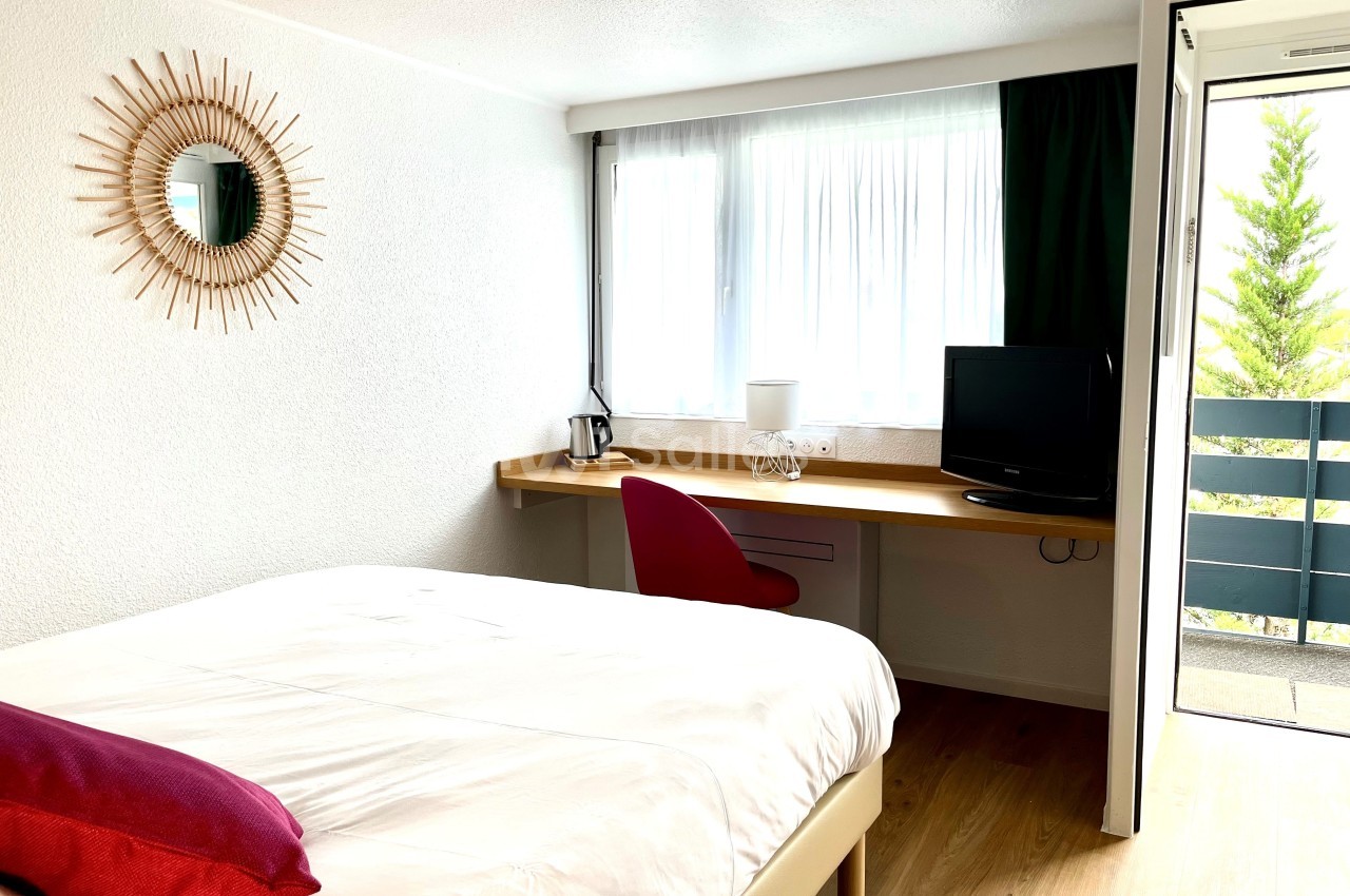 Chambre lumineuse avec lit, bureau équipé d'une chaise rouge, télévision et balcon donnant sur l'extérieur.