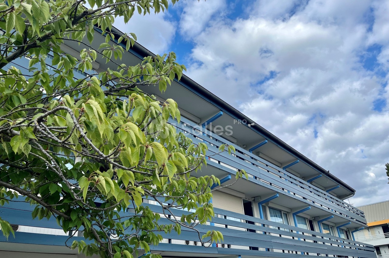 Façade d'un bâtiment à étages avec balcons bleus, partiellement cachée par un arbre sous un ciel nuageux.