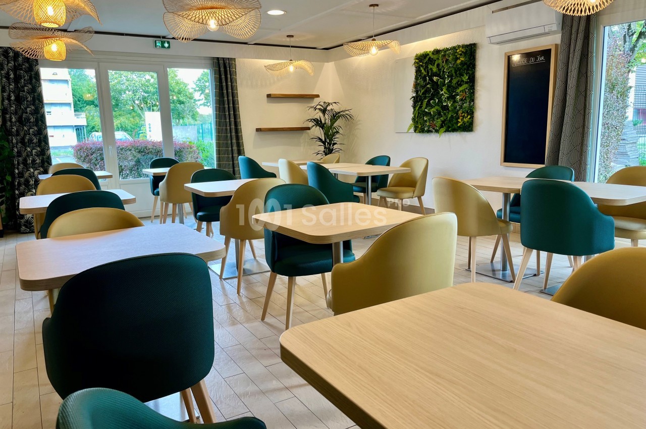 Salle lumineuse avec tables en bois clair, chaises colorées et mur végétal, donnant sur un extérieur verdoyant.