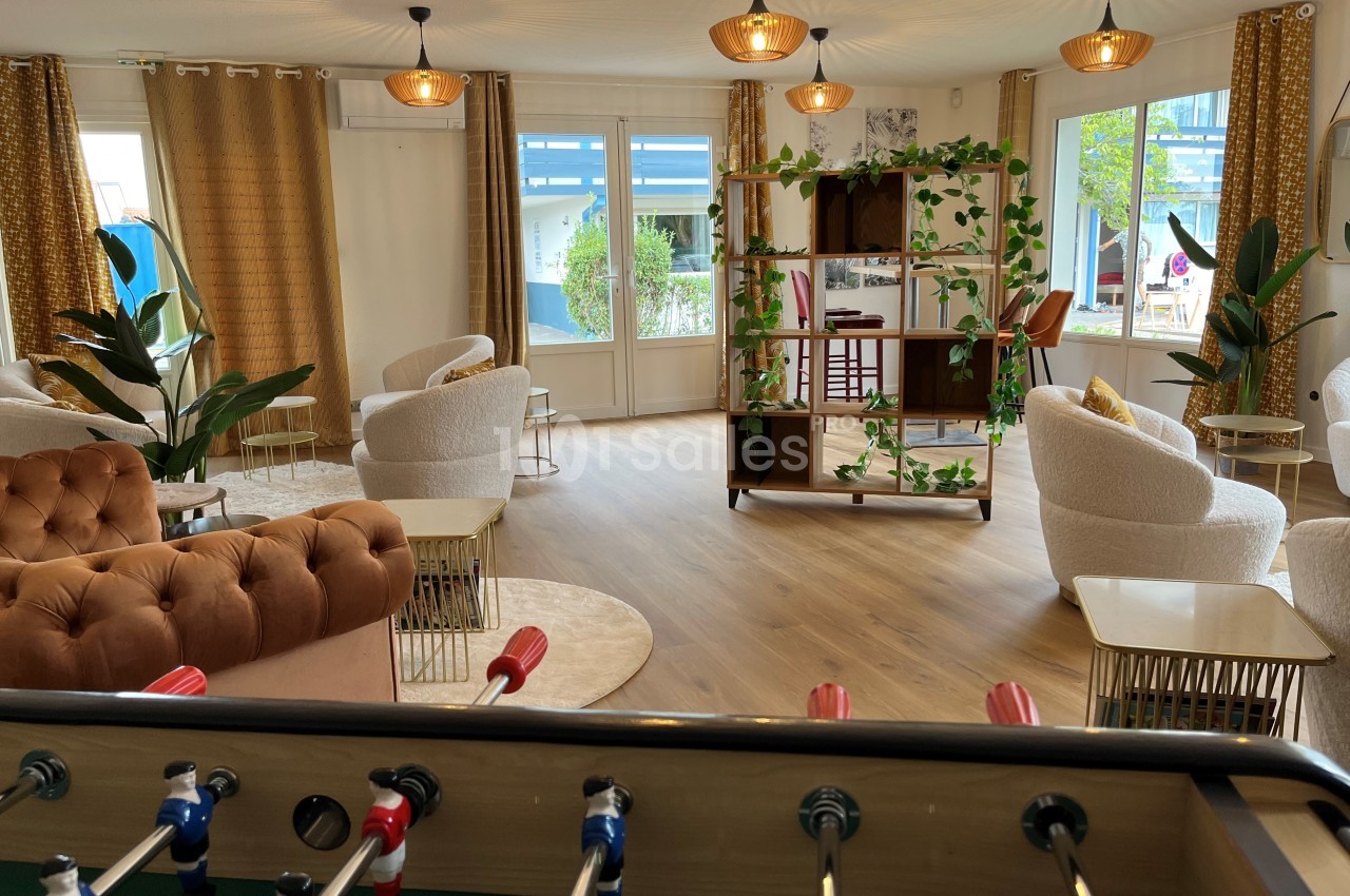 Espace lumineux avec fauteuils, étagères décorées, baby-foot au premier plan et grandes fenêtres donnant sur l'extérieur.