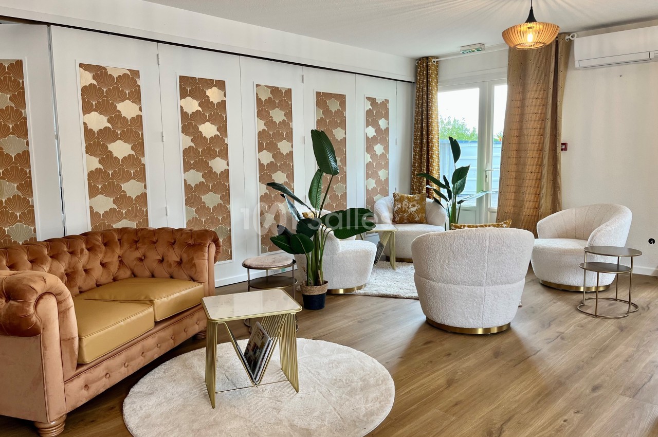 Salon lumineux avec canapé en cuir, fauteuils blancs, plantes vertes et table basse sur un parquet en bois.