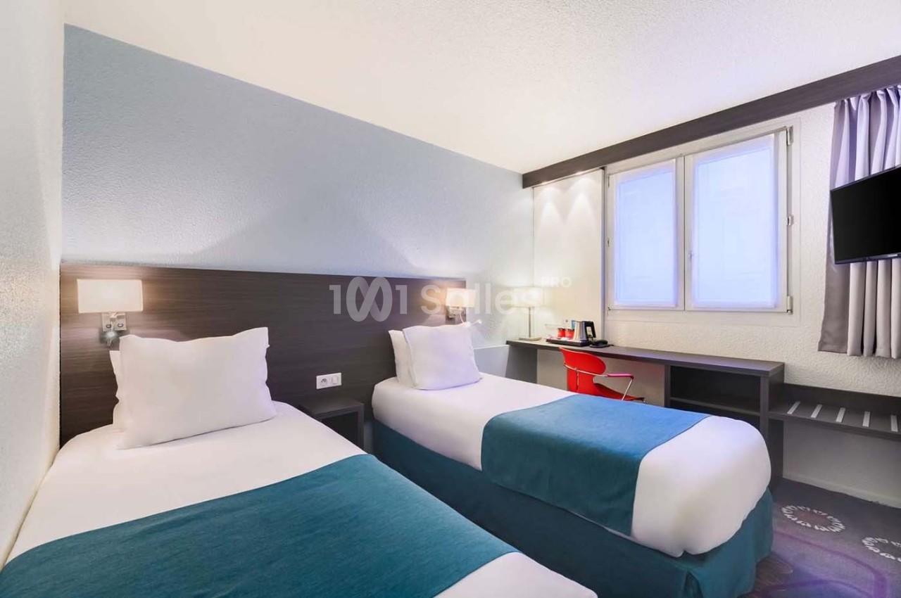 Chambre avec deux lits simples, bureau, chaise rouge, télévision murale et fenêtre laissant entrer la lumière naturelle.