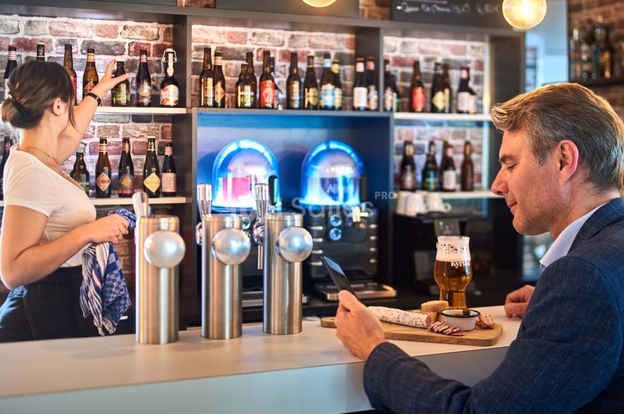 Un homme au comptoir d'un bar lit sur son téléphone tandis qu'une serveuse range des bouteilles sur une étagère.