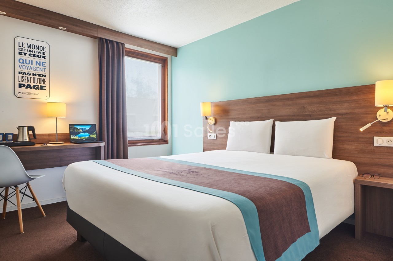 Chambre d'hôtel avec lit double, bureau, chaise, lampe et mur turquoise, éclairée par une fenêtre.