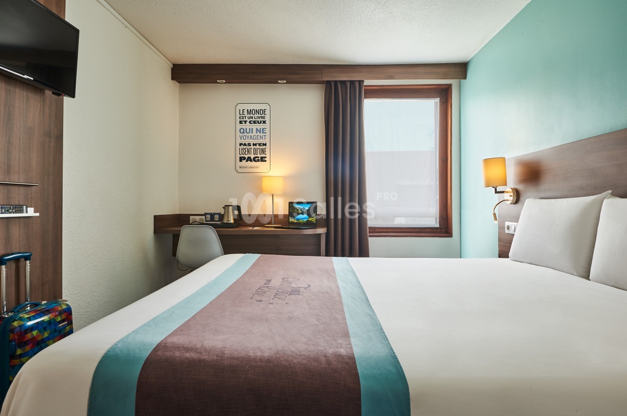 Chambre d'hôtel avec lit double, bureau, lampe allumée, valise colorée et fenêtre avec rideau partiellement ouvert.
