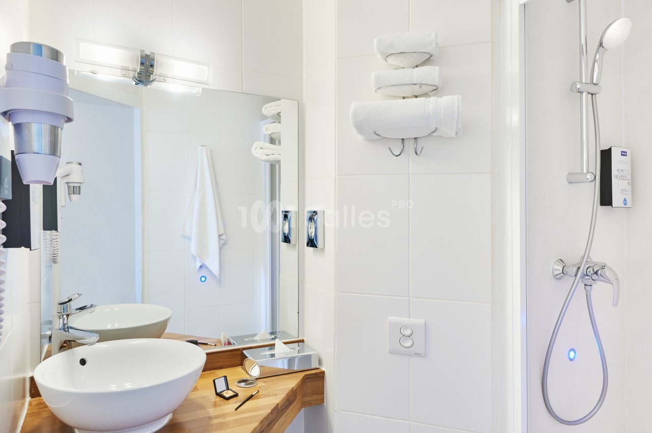 Salle de bain moderne avec lavabo, miroir, sèche-cheveux, serviettes pliées et douche équipée d'une pomme de douche.