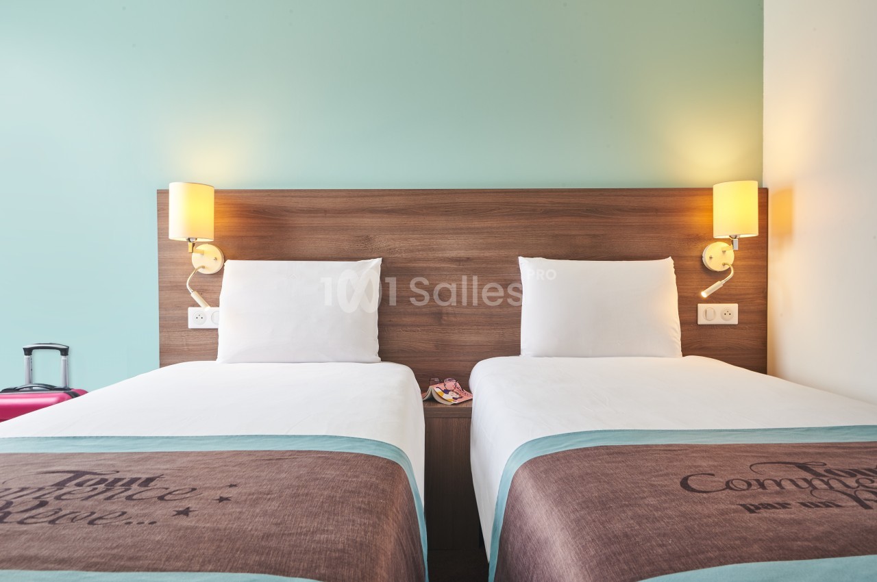 Deux lits simples avec tête de lit en bois, lampes murales allumées et mur bleu clair en arrière-plan.